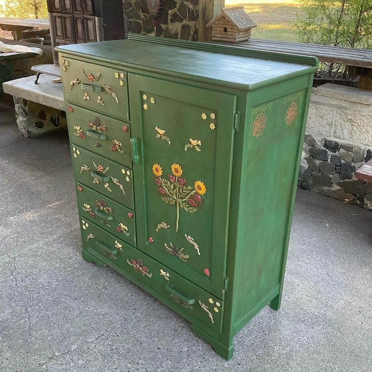 a green cabinet with flowers painted on the doors and drawers is sitting in front of a bench