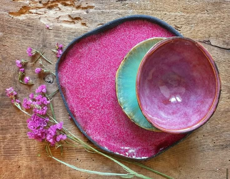 two pink and green bowls sitting on top of a wooden table next to purple flowers