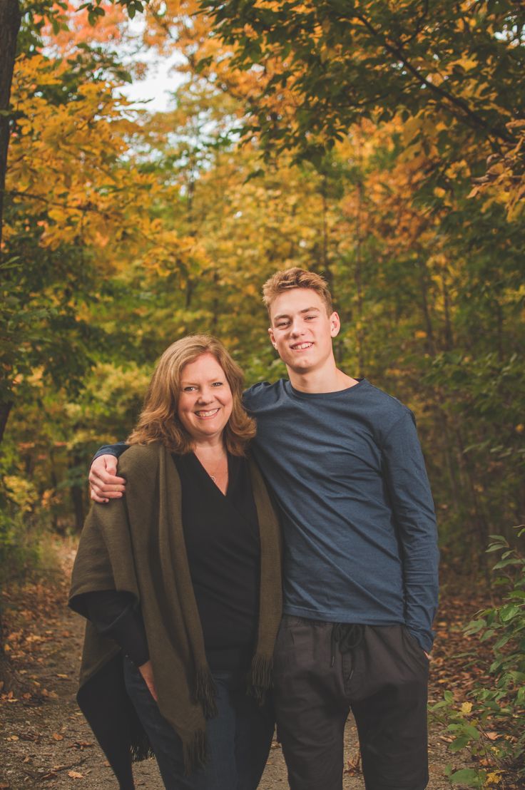 a man and woman posing for a photo in front of trees with fall leaves on the ground