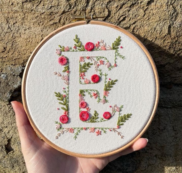 a person holding up a cross stitched monogram in front of a stone wall