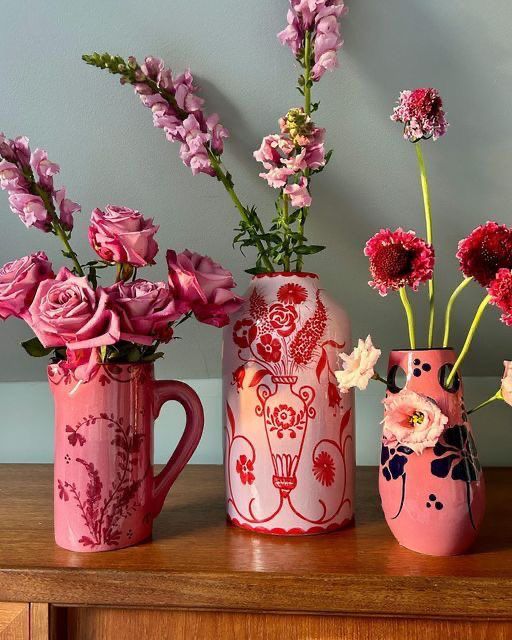 three pink vases with flowers in them on a table next to a wall and a wooden cabinet