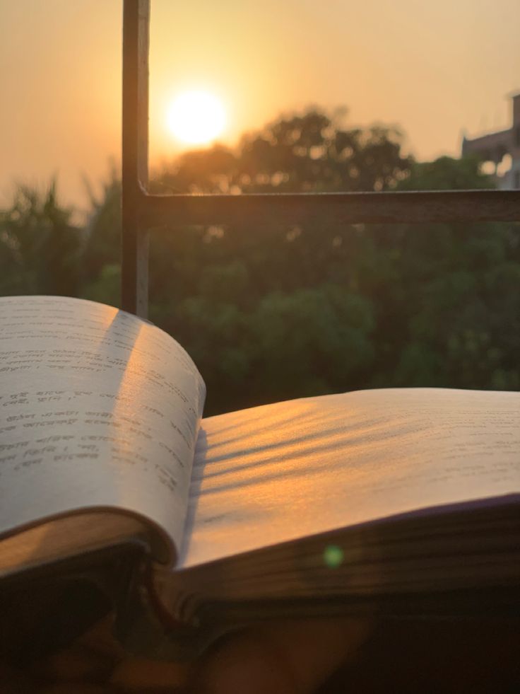 an open book sitting on top of a table next to a window with the sun setting