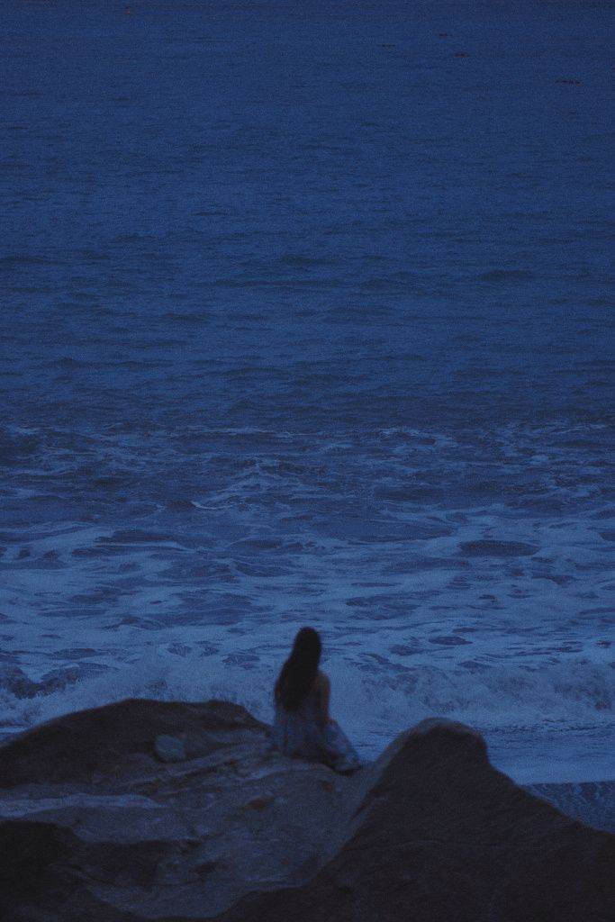 a woman sitting on top of a rock next to the ocean at night with an umbrella over her head