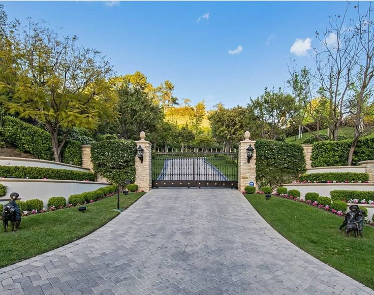 the driveway is lined with hedges and stone pillars, leading to a gated entrance