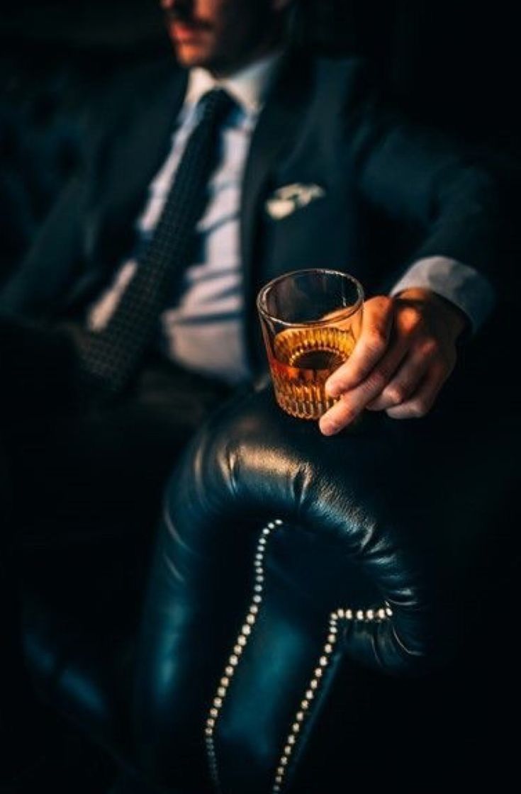 a man sitting in a chair holding a glass with something on the arm and wearing a tie