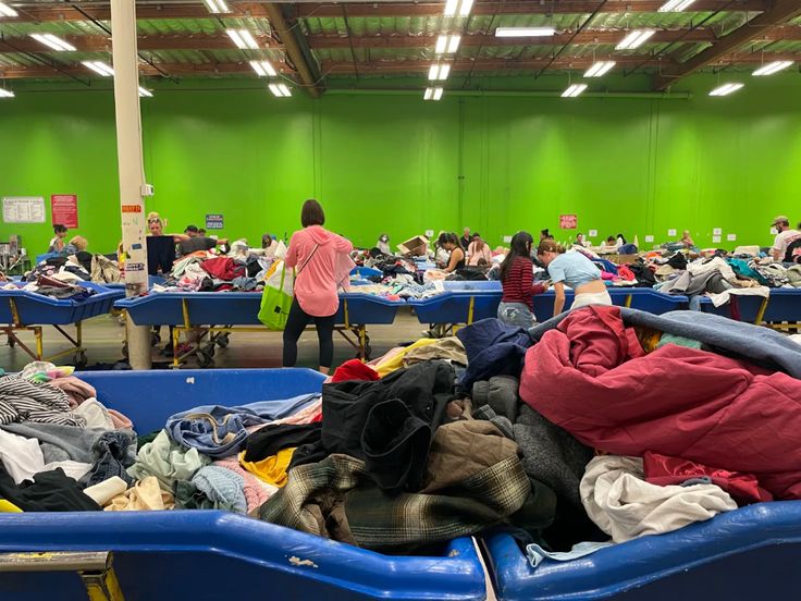 many people are standing around in a room full of clothes and other items on tables