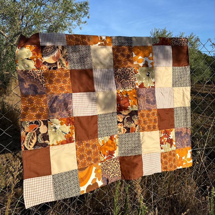 a patchwork quilt hanging on a chain link fence with trees in the back ground