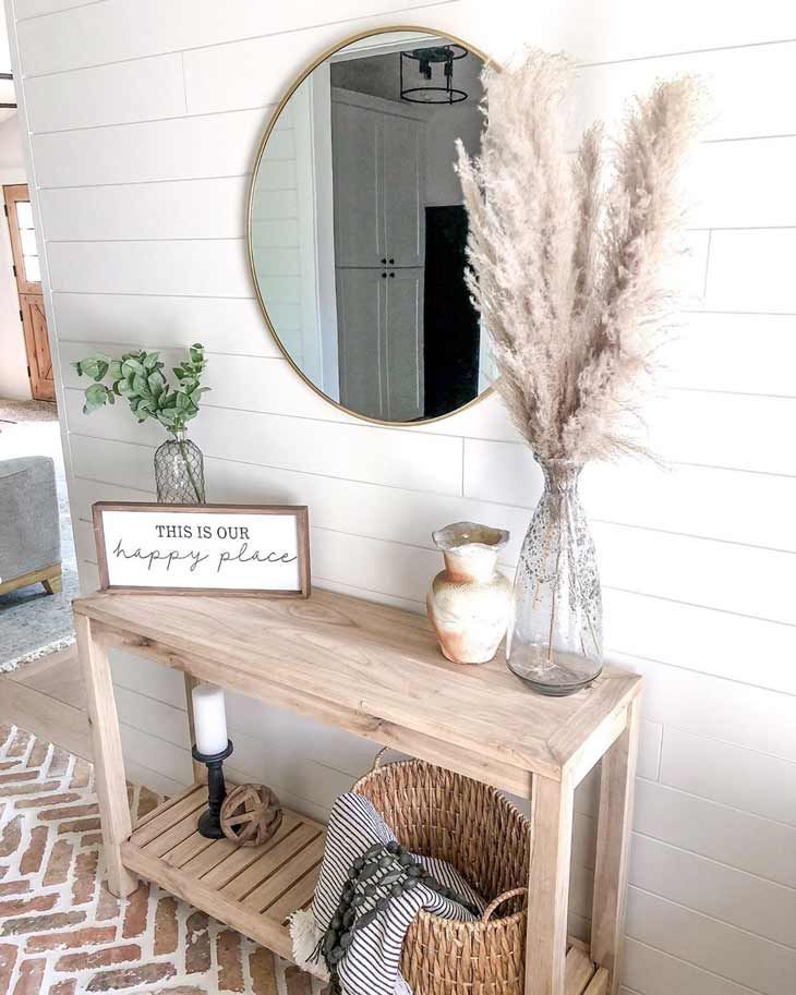 a wooden table with a mirror, vase and some plants on it in front of a white wall