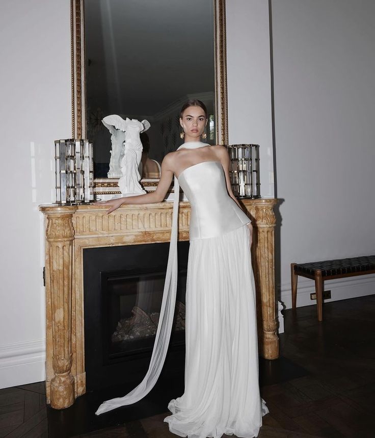 a woman standing in front of a fireplace wearing a white dress and holding a long veil