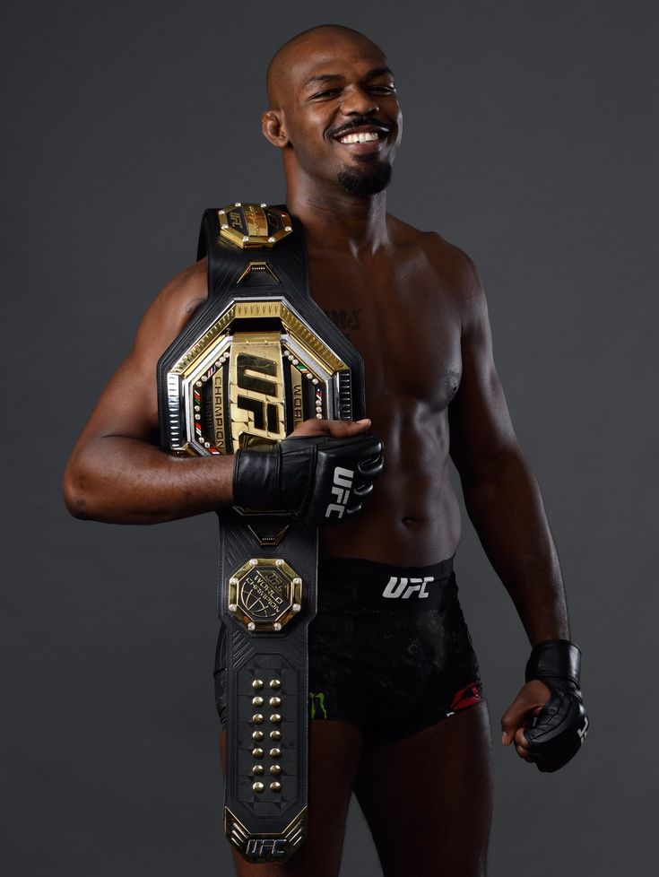 a man holding a wrestling belt in his right hand and smiling while standing against a gray background