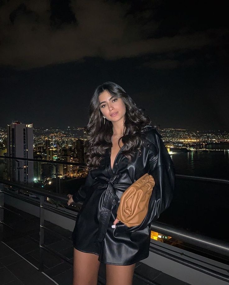 a woman standing on top of a roof next to a city at night with her hands in her pockets