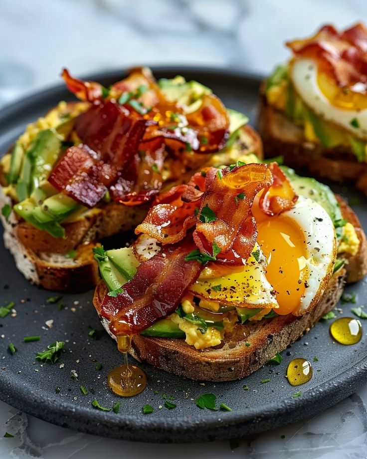 an open face sandwich with bacon, eggs and avocado on a black plate