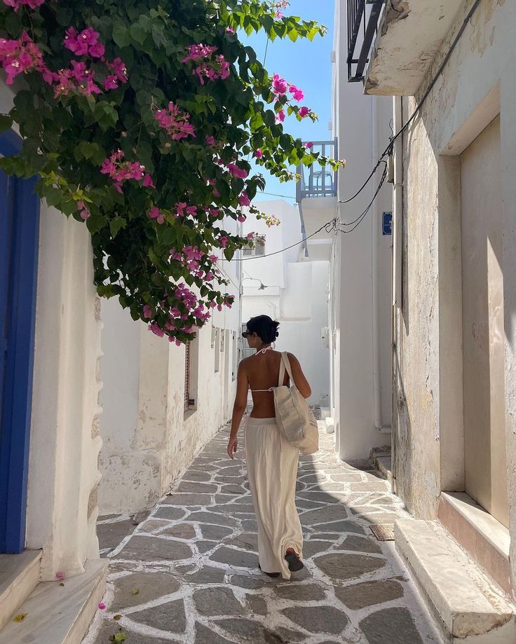 a woman walking down an alley way with pink flowers on the trees above her head