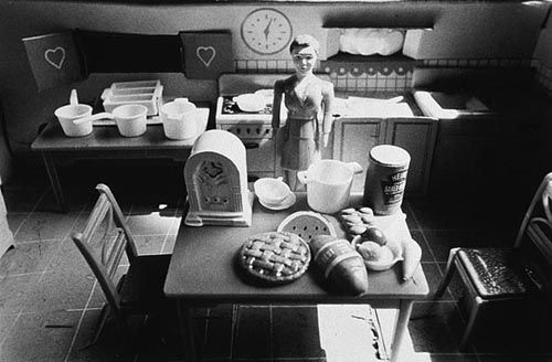 a black and white photo of a woman in the kitchen with food on the table