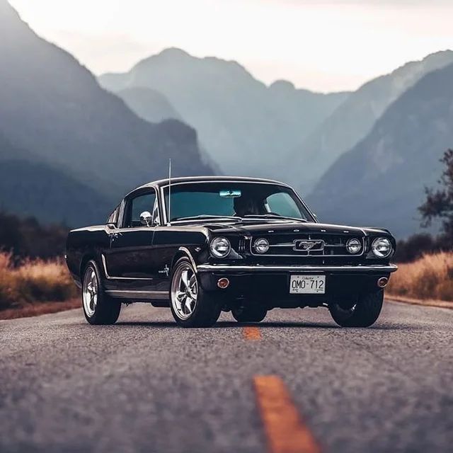 an old black car is parked on the side of the road with mountains in the background