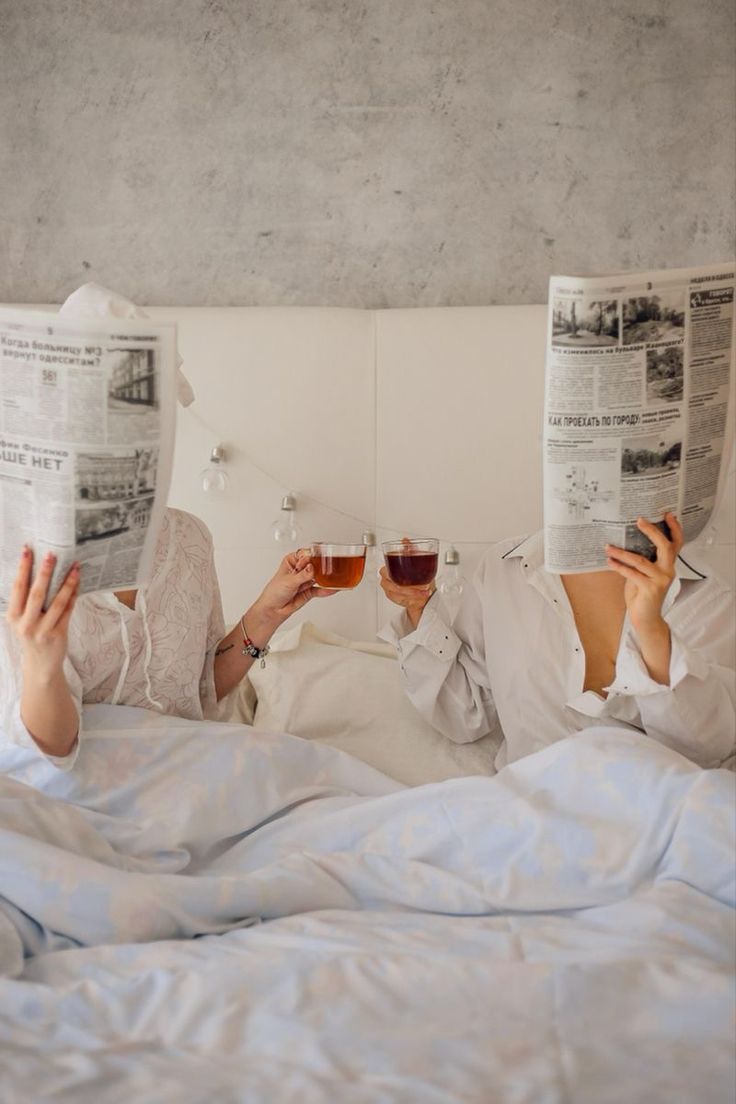 two women laying in bed reading newspapers and drinking red wine while holding glasses of wine