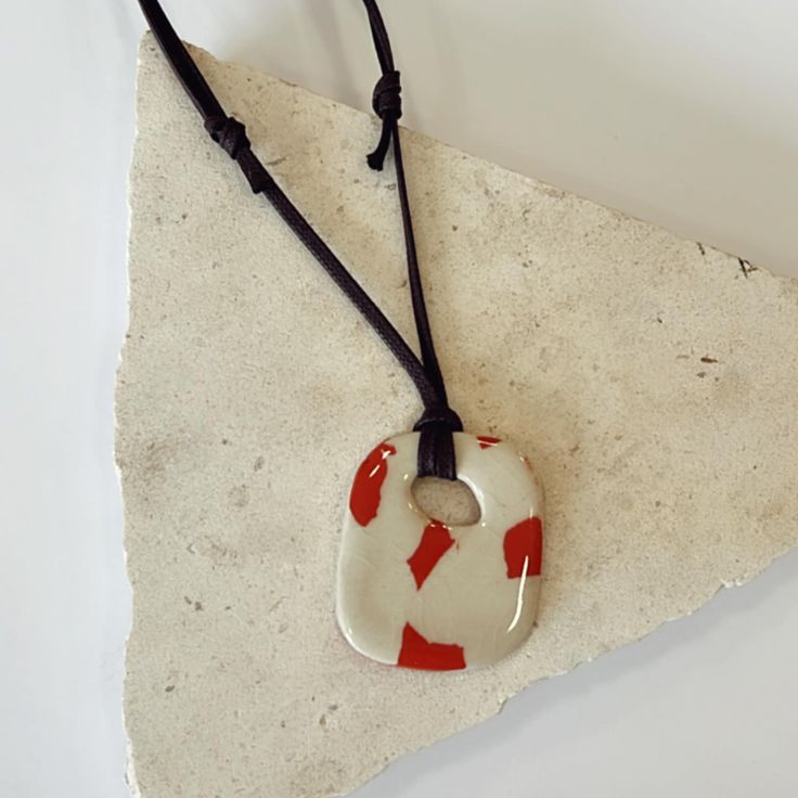 a red and white ceramic pendant hanging on a black leather cord with a square hole in the middle