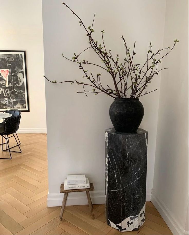 a black vase with branches in it sitting next to a white wall and wooden floor