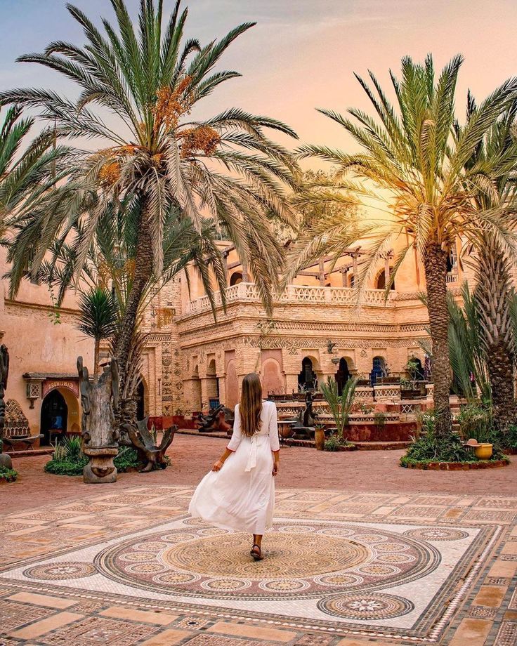 a woman in a white dress is walking through a courtyard surrounded by palm trees and potted plants