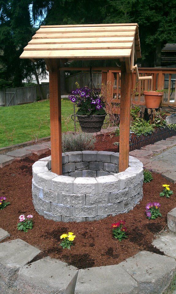 a stone fire pit surrounded by flowers and plants in a backyard area with a wooden gazebo