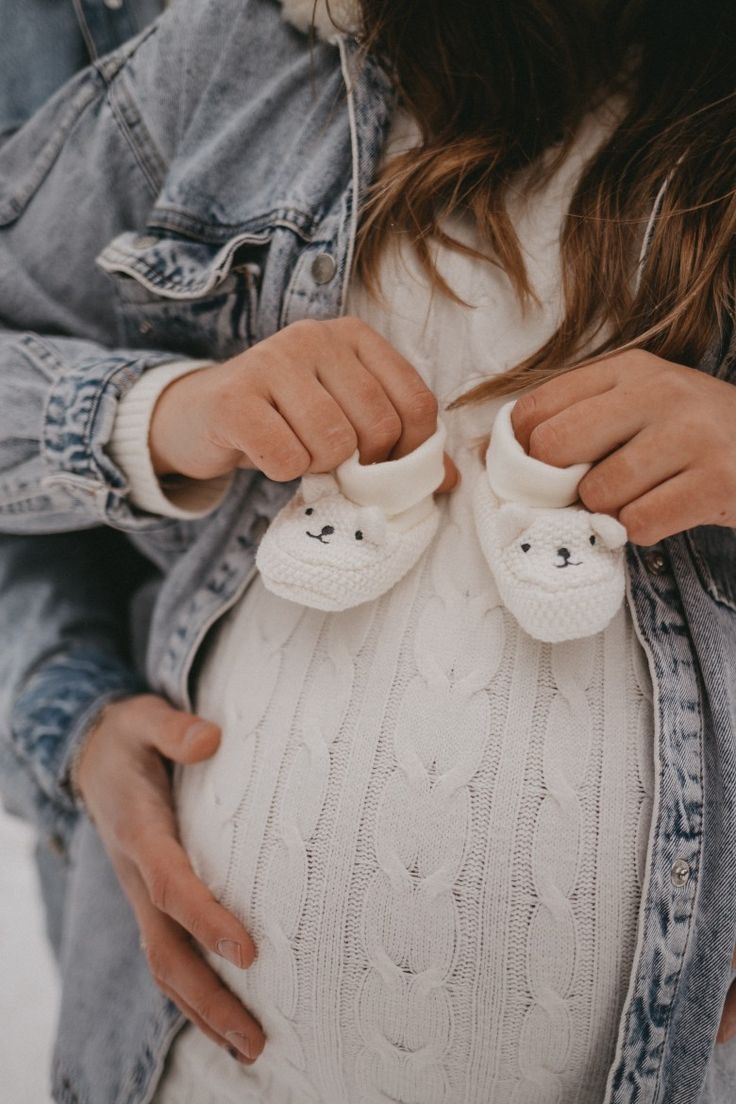 a pregnant woman is holding her shoes while wearing a white sweater and jean jacket with bear buttons