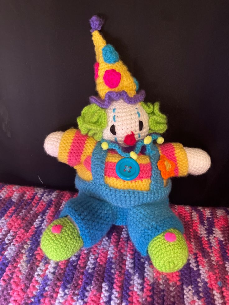 a crocheted clown stuffed animal sitting on top of a purple and pink blanket