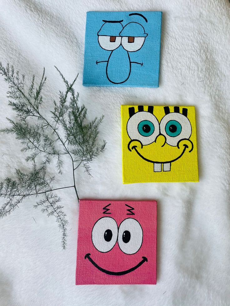 three square paintings with faces on them sitting next to a small green plant and white towel