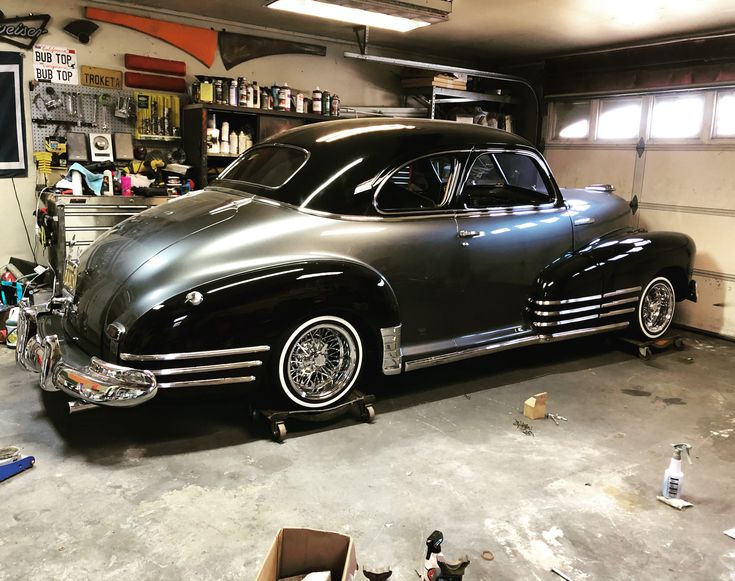 an old black car in a garage with tools on the floor and other items around it