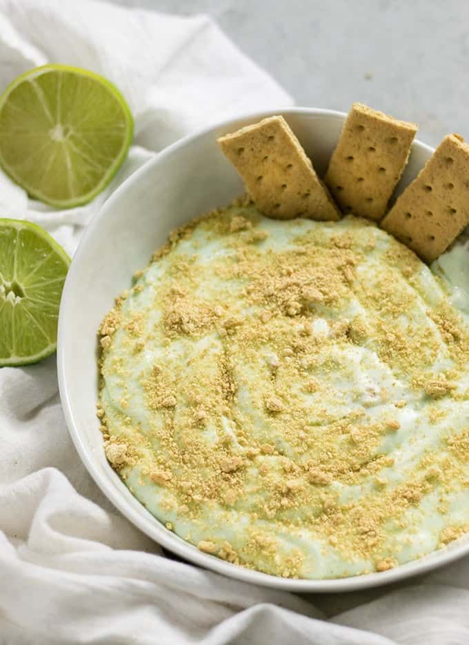 a white bowl filled with green dip and crackers next to limes on the side