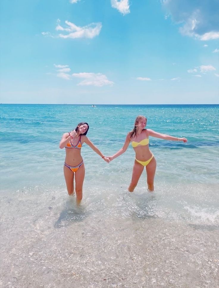 two women in bikinis holding hands while standing in the water at the beach on a sunny day