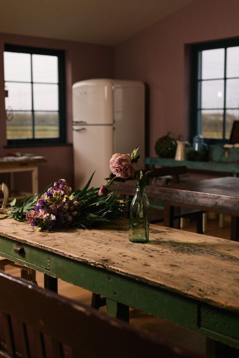 flowers sit on top of a wooden table in a room with two windows and a refrigerator