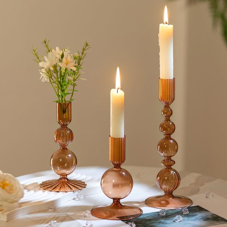 three candles are sitting on a table with flowers in vases next to them,