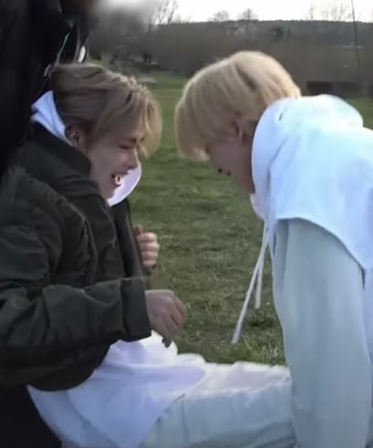 two people sitting on the ground with one person holding something in his mouth and another standing behind them