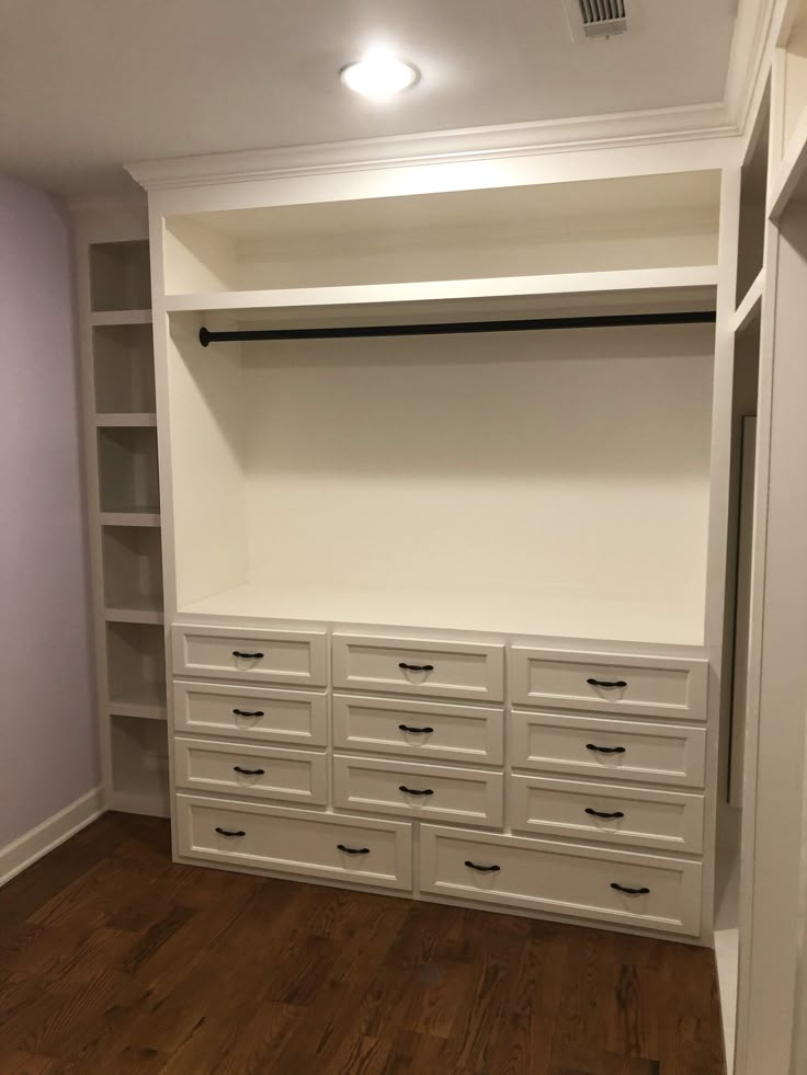 an empty walk in closet with white cabinets and drawers on the wall, along with wooden flooring