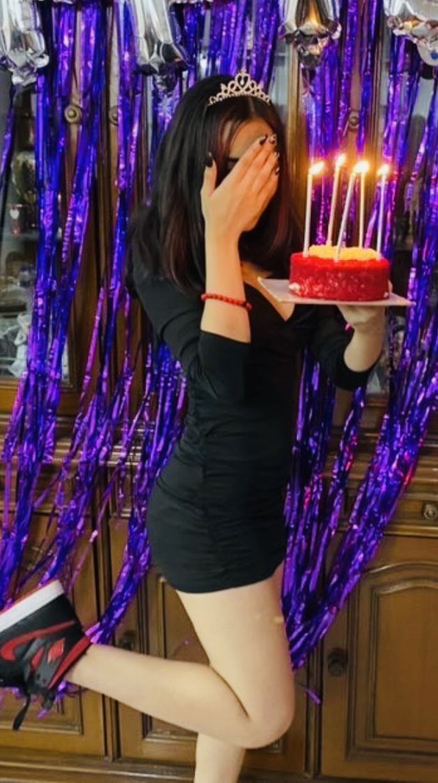 a woman holding a cake with lit candles on it in front of purple streamers