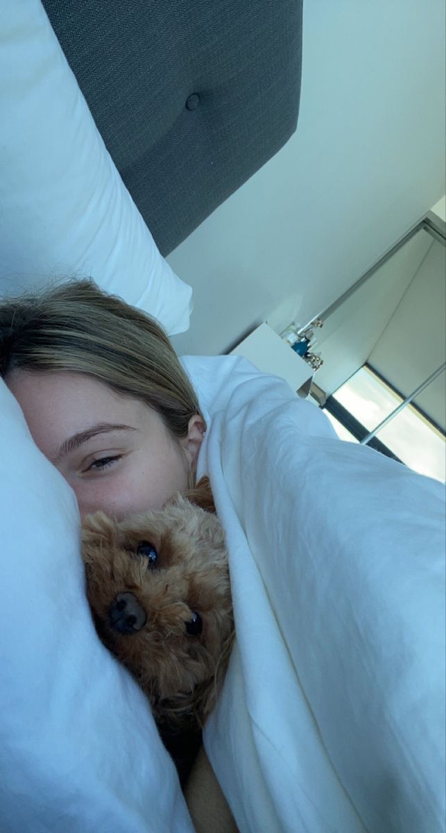 a woman laying in bed with her head on the pillow while holding a small dog