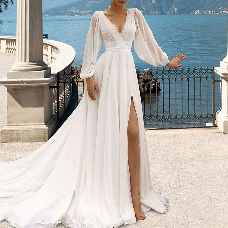 a woman standing in front of a lake wearing a white dress with long sleeves and high slit