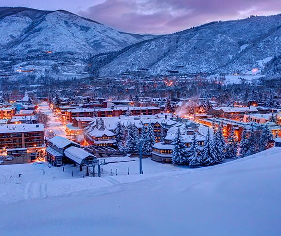 the town is lit up at night in the snow covered mountain area with mountains behind it