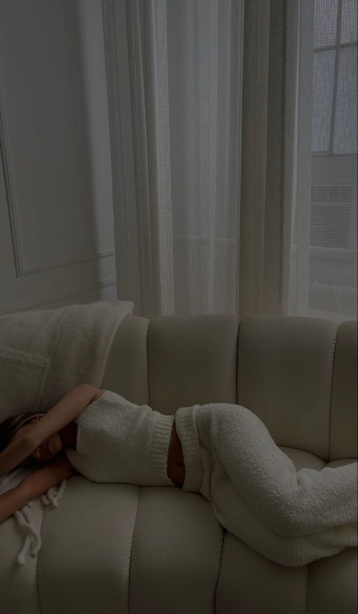 a woman laying on top of a couch covered in white blanket next to a window