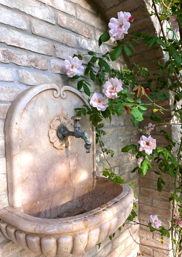 a water fountain with pink flowers growing on the side of it next to a brick wall