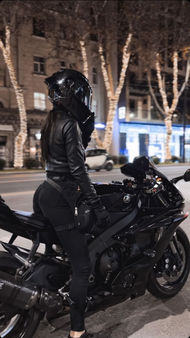 a woman in black riding on the back of a motorcycle at night with trees and buildings behind her