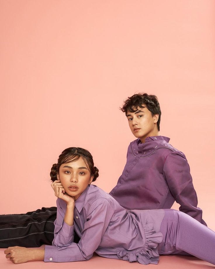 two young people sitting on the ground posing for a photo in front of a pink background