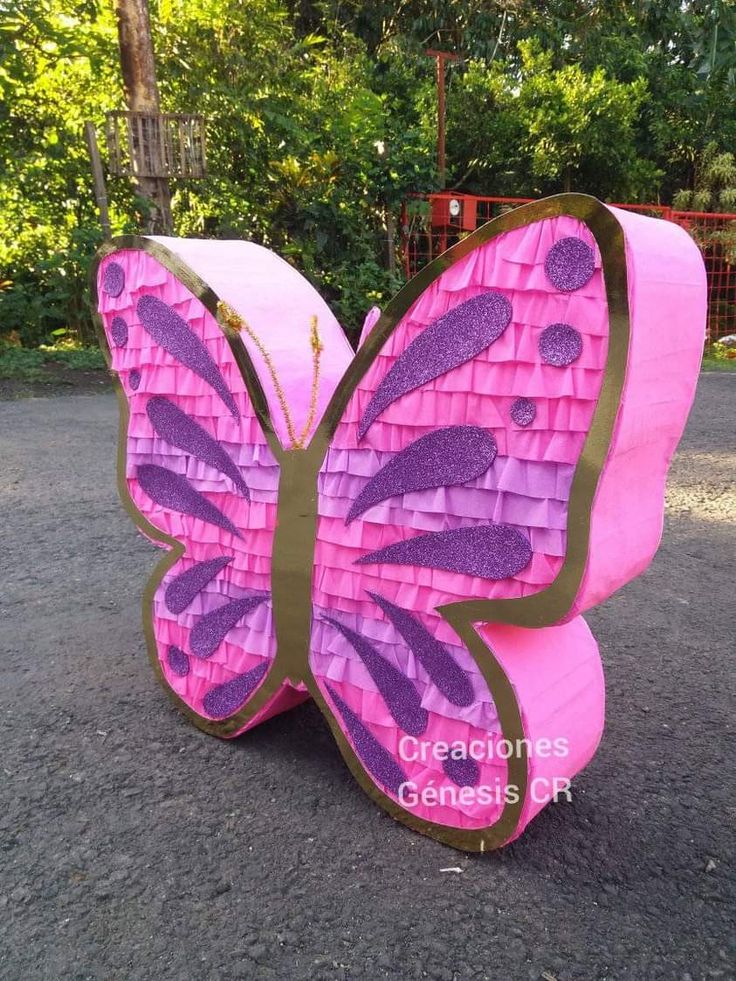 a pink and purple butterfly sculpture sitting on top of a gravel ground next to trees