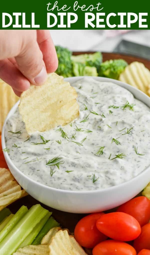 a person dipping dip into a white bowl with chips and vegetables around it on a platter