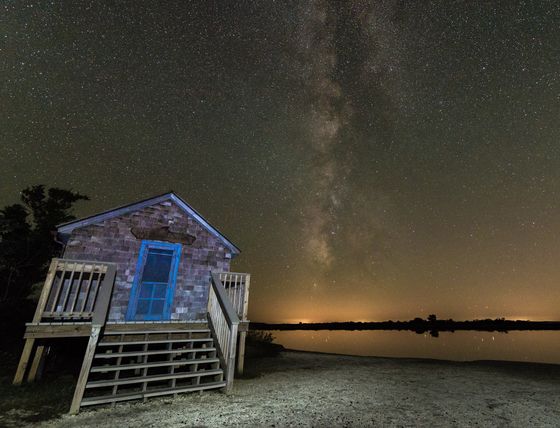 the night sky is filled with stars, and there are two small buildings on the shore