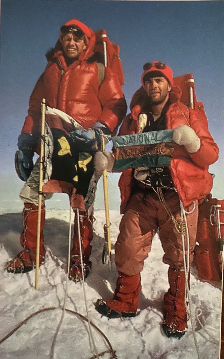 two men in ski gear standing on top of a snow covered slope holding skis