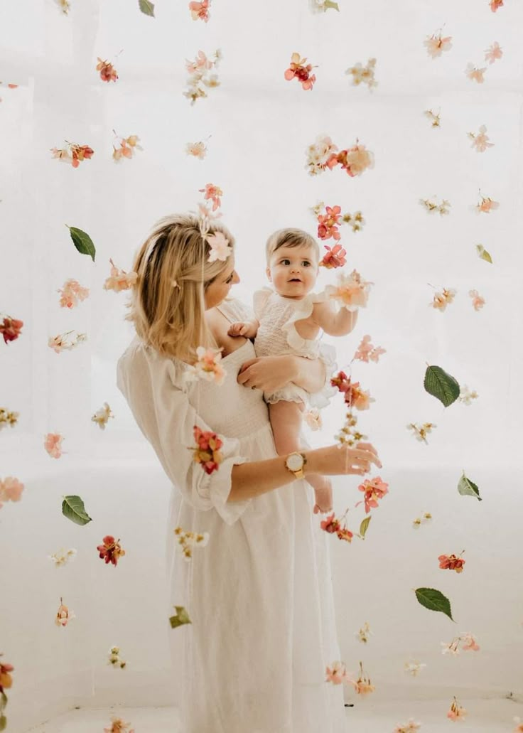 a woman holding a baby while standing in front of a floral wall with flowers on it