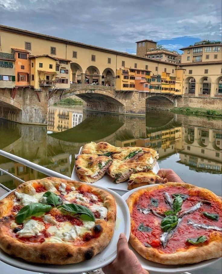 two pizzas are sitting on plates in front of a river with buildings and bridges