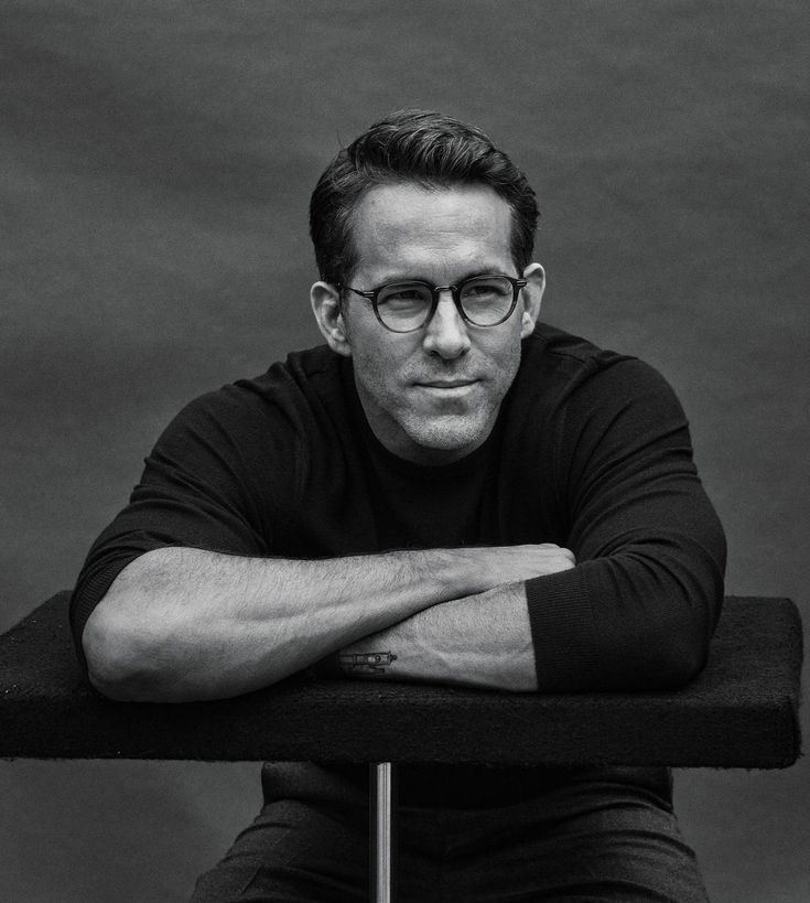 a black and white photo of a man with glasses sitting on a chair looking at the camera