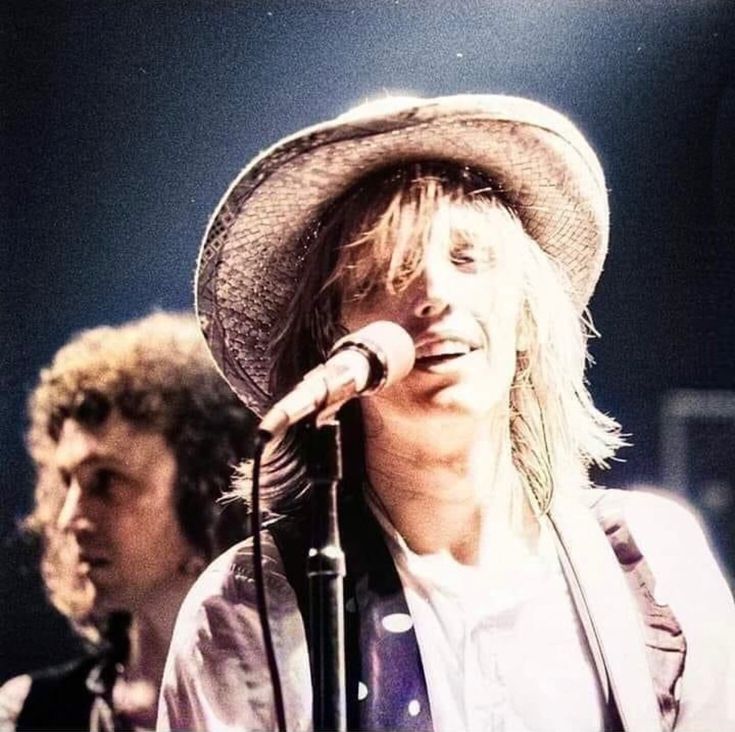 a man in a hat singing into a microphone while another man plays the guitar behind him